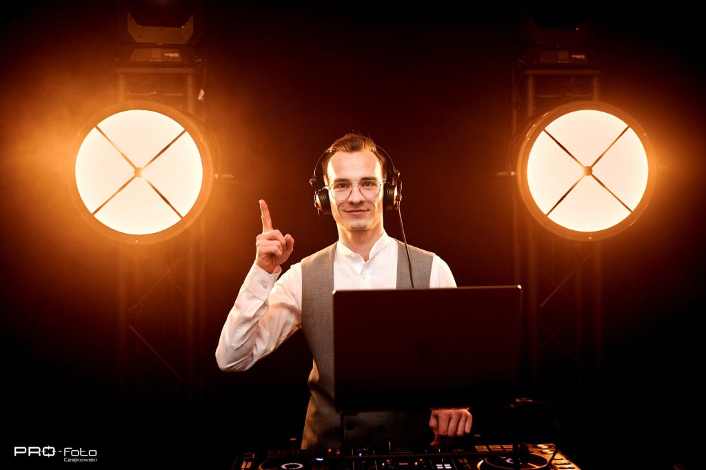 DJ Fuller w słuchawkach, z podniesioną ręką, między dwiema świecącymi lampami.
