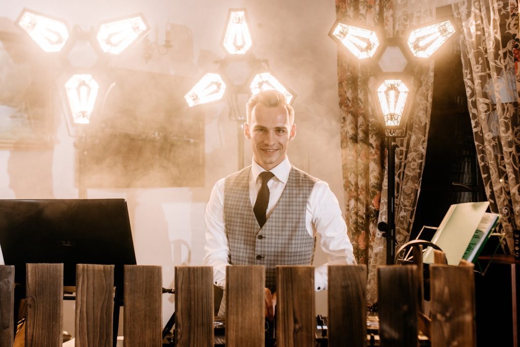 DJ Fuller za drewnianą konsoletą, w tle ciepłe żarówki i lekki dym.